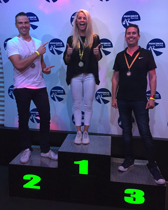 the winner giving two thumb's up as she stands on the winner's box, while the second place position points and smiles at her